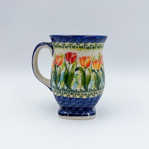 May include: A ceramic mug with a floral tulip design. The mug features a blue rim and base, with a cream-colored body adorned with red, orange, and green tulips. The handle is a light beige color.