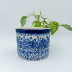 May include: A ceramic planter with a blue floral pattern. The planter has a dark blue rim and a white interior. The floral design features various blue flowers and foliage. The planter is cylindrical and sits on a white surface.