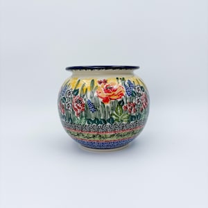 May include: A round ceramic vase with a floral design. The vase features a vibrant array of red, yellow, blue, and green flowers and foliage. The rim is dark blue, and the base has a decorative border.