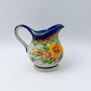 May include: A ceramic creamer pitcher featuring a cream-colored base with a hand-painted floral pattern in shades of yellow, orange, pink, and green. The rim and handle are accented with a deep blue glaze, and the body has a rounded shape.