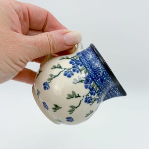 May include: A small, ceramic creamer with a handle, featuring a floral design in blue and green on a cream background. The rim is decorated with a blue and white pattern. The creamer is held in a hand.