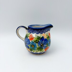 May include: A ceramic creamer pitcher featuring a hand-painted floral pattern with blue and orange flowers, green leaves, and a dark blue circular border design around the rim. The pitcher has a curved handle and a small pouring spout.