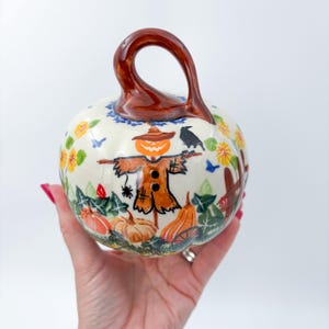 May include: A ceramic pumpkin-shaped decorative piece with a glossy brown stem handle. The cream-colored surface features hand-painted illustrations of a scarecrow, a jack-o'-lantern, a black crow, orange pumpkins, green leaves, and yellow flowers.