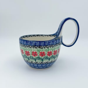 May include: A ceramic soup bowl with a large handle. The bowl features a floral pattern with red flowers and green leaves on a white background. The rim and handle are blue, with a decorative border.