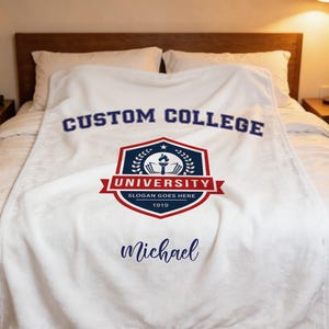 Könnte beinhalten: Eine weiße Fleecedecke mit dem dunkelblauen Schriftzug CUSTOM COLLEGE oben. Darunter befindet sich ein rot-blaues Wappen mit einer Fackel, einem Buch und dem Text UNIVERSITY, SLOGAN GOES HERE sowie 1919. Der Name Michael ist in Blau gedruckt.