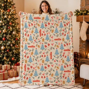 May include: A cream-colored fleece blanket featuring a repeating Christmas pattern of light blue pine trees, brown gingerbread men, red mittens, and green holly leaves, with the name emilia printed in red text throughout the design.