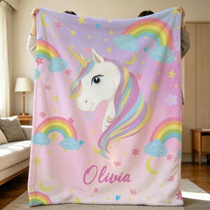 May include: A soft fleece blanket featuring a central white unicorn with a multicolored mane and horn. The background is light pink and purple, decorated with rainbows, blue clouds, yellow stars, crescent moons, and flowers. The name Olivia is printed.