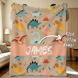 Op de afbeelding: Een beige deken met een herhalend patroon van kleurrijke dinosaurussen, vulkanen en planten. Een horizontale perzikkleurige band in het midden toont de naam JAMES in witte letters. Een pijl wijst naar de tekst YOUR CUSTOM NAME.