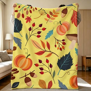 Könnte beinhalten: Eine hellgelbe Fleecedecke mit einem sich wiederholenden Muster aus herbstlichen Illustrationen, darunter orangefarbene Kürbisse, braune Eicheln, rote Beeren an dünnen Stielen sowie verschiedene bunte Blätter in Rot, Blau und Dunkelgrün.