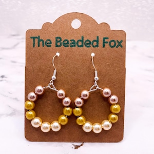 May include: A pair of hoop earrings with a mix of yellow, peach, and white beads. The earrings are displayed on a brown card with the text "The Beaded Fox" in green. The earrings have silver hooks.