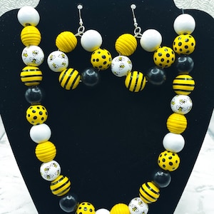 May include: A necklace and earring set featuring large, round beads in yellow, white, and black. Some beads have bee designs, stripes, or polka dots. The jewelry is displayed on a black stand.