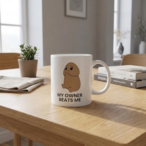 以下が含まれることがあります： 悲しそうな茶色のキャラクターの漫画イラストが描かれた白いセラミックマグ。テキスト「MY OWNER BEATS ME」が下に黒で印刷されています。マグカップには湾曲したハンドルが付いています。