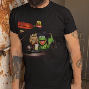 May include: A black short-sleeved t-shirt featuring a graphic print of Miss Piggy and Kermit the Frog sitting in a car at a McDonald's drive-thru. Kermit is shown with a hand gesture, and the McDonald's logo is visible above the car roof.