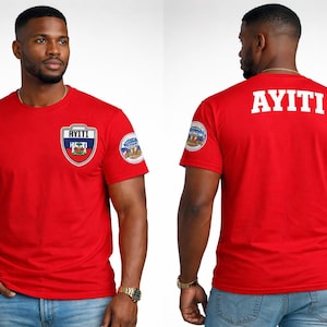 Puede incluir: Camiseta roja con la palabra "AYITI" en blanco en la espalda. La parte delantera presenta un escudo con la bandera haitiana y la palabra "AYITI". Pequeños parches circulares están en las mangas.