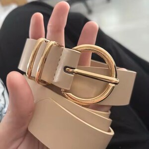 May include: A beige belt featuring a smooth, solid-coloured strap and a polished gold-tone circular buckle. The belt includes two matching gold-tone metal loops positioned near the buckle, providing a secure and structured look for this accessory.