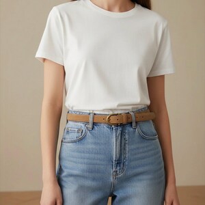 May include: A plain white short-sleeved t-shirt tucked into light blue denim jeans. A tan suede belt with a brass-toned metal buckle is threaded through the belt loops of the jeans, cinching the waistline of the garment ensemble.