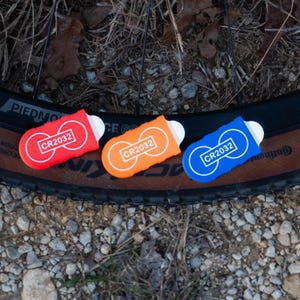 May include: Three colorful CR2032 battery holders in red, orange, and blue, each with a white label and the text "CR2032". The holders are placed on a bicycle tire, with a gravel background.