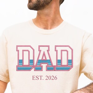 May include: Cream-colored t-shirt with the word "DAD" in a pink and blue retro design. Below the word "DAD" is the text "EST. 2026". The shirt is made of a soft material.