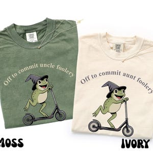 May include: Two t-shirts, one moss green and one ivory, each with a graphic of a frog riding a scooter and wearing a witch hat. The text on the moss green shirt reads "Off to commit uncle foolery." The ivory shirt says "Off to commit aunt foolery."
