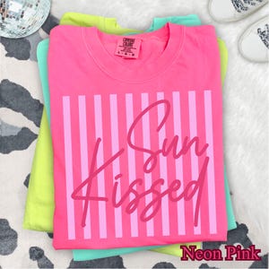 May include: A stack of three Comfort Colors t-shirts in neon colors. The top shirt is neon pink with vertical stripes and the words "Sun Kissed" in a script font. The other shirts are light green and teal. The bottom right corner of the image has the words "Neon Pink".