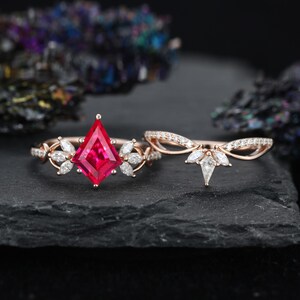May include: A rose gold ring set featuring a kite-shaped red center stone flanked by marquise-cut white stones, paired with a matching curved band adorned with small white stones and a central kite-shaped white stone on a dark textured surface.