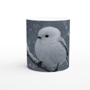 以下が含まれることがあります： 雪の降る背景に、雪に覆われた枝に止まっているふわふわの白い鳥が描かれた白いセラミックマグ。鳥は黒い目と小さなくちばしをしています。マグカップは円筒形で、白いハンドルが付いています。
