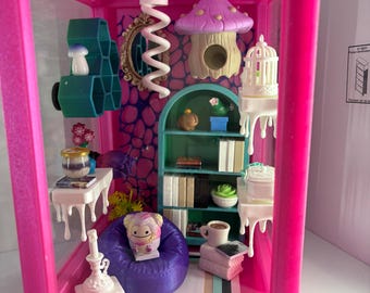 Può includere: Una stanza in miniatura all'interno di una cornice rosa brillante con un pouf viola, una libreria verde acqua con libri, un tavolo bianco con un barattolo, una luce a spirale bianca sospesa, una casa a fungo viola e una piccola statuetta.