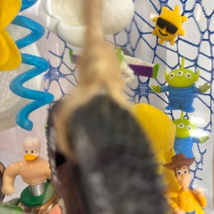 May include: A clear display case containing various small plastic character figurines including a cowboy, green aliens, a yellow sun with sunglasses, a white cloud, a blue spiral, and a duck figure, with a tan rope hanging in the foreground.