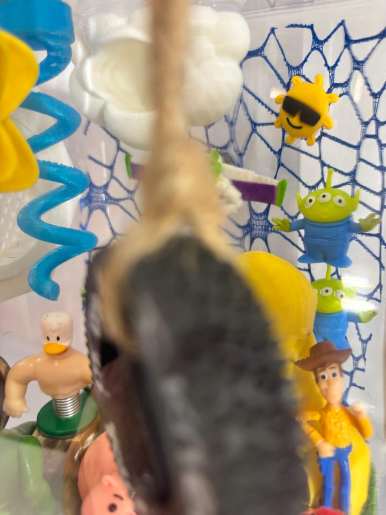 May include: A clear display case containing various small plastic character figurines including a cowboy, green aliens, a yellow sun with sunglasses, a white cloud, a blue spiral, and a duck figure, with a tan rope hanging in the foreground.
