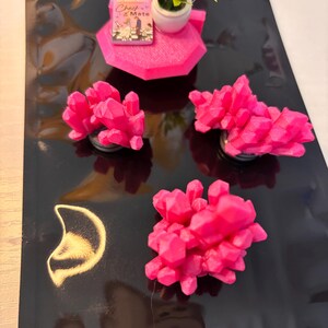 Könnte beinhalten: Eine Sammlung von rosa kristallartigen Skulpturen, eine kleine rosa Plattform mit einer Miniaturpflanze in einem weißen Topf und ein Buch mit dem Titel "Check & Mate" auf einer schwarzen Oberfläche. Die Kristalle haben ein facettiertes Design.