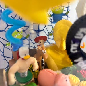 May include: A collection of small plastic character figurines including a duck with a spring base, a brown horse, a cowgirl, a pink pig, and two green three-eyed aliens. A large yellow bust and a blue spiral shape are also visible in the display.