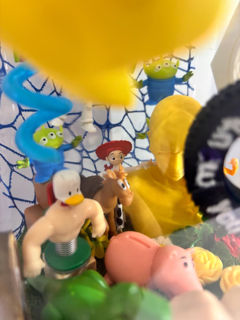 May include: A collection of small plastic character figurines including a duck with a spring base, a brown horse, a cowgirl, a pink pig, and two green three-eyed aliens. A large yellow bust and a blue spiral shape are also visible in the display.