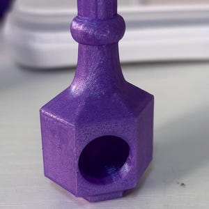 Puede incluir: Un tapón de botella morado, impreso en 3D, con base hexagonal y parte superior cilíndrica. El tapón tiene una superficie texturizada y una abertura circular en un lado. El diseño es simple y moderno.