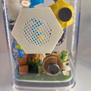 May include: A clear rectangular container housing a miniature diorama featuring a white hexagonal speaker grille, a yellow flower, a blue spiral component, and small green figures on a green base. The structure includes various small objects.