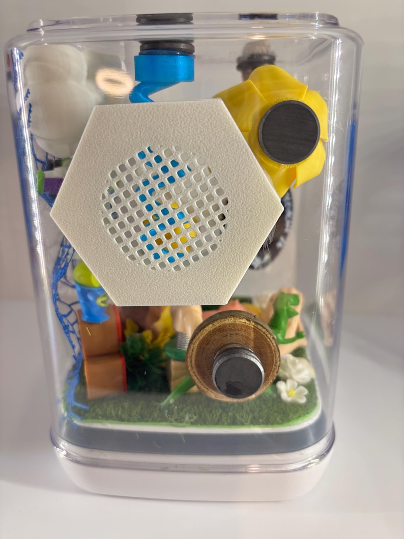 May include: A clear rectangular container housing a miniature diorama featuring a white hexagonal speaker grille, a yellow flower, a blue spiral component, and small green figures on a green base. The structure includes various small objects.