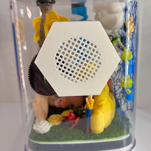 May include: A clear rectangular display case containing miniature figures including a yellow star, a brown tire swing, a pink pig, and small green aliens on a blue net. A white hexagonal speaker grille is mounted on the front of the clear plastic casing.