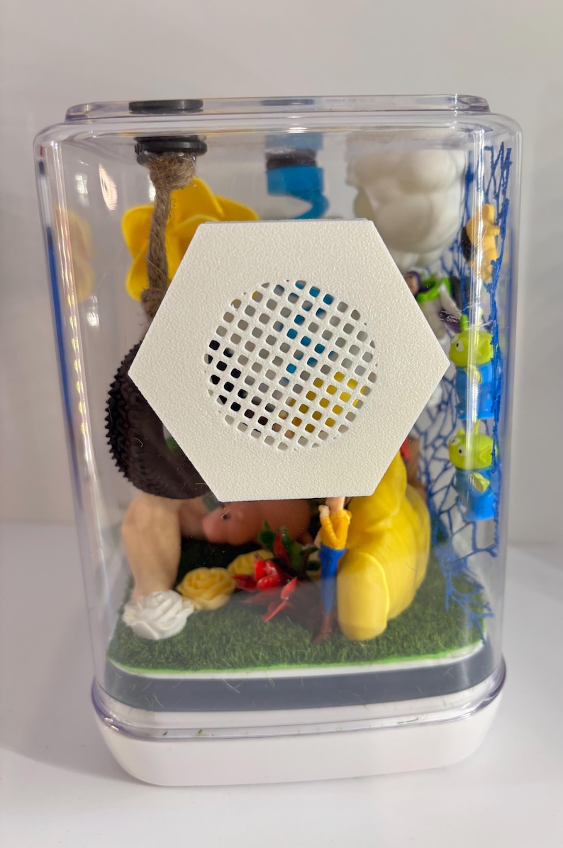 May include: A clear rectangular display case containing miniature figures including a yellow star, a brown tire swing, a pink pig, and small green aliens on a blue net. A white hexagonal speaker grille is mounted on the front of the clear plastic casing.