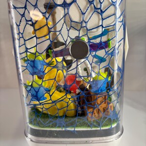 May include: A clear rectangular display case containing a collection of small plastic character figurines, including green aliens and a space ranger figure, arranged on a green base. The front of the case is covered by a blue web-like pattern.