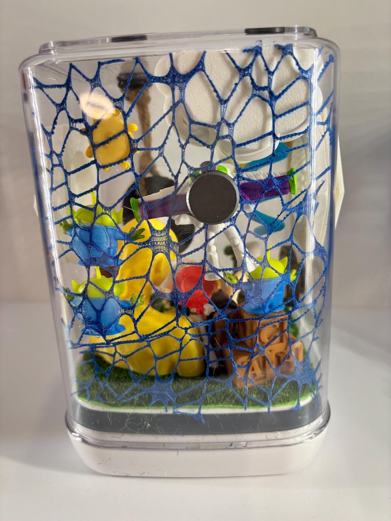 May include: A clear rectangular display case containing a collection of small plastic character figurines, including green aliens and a space ranger figure, arranged on a green base. The front of the case is covered by a blue web-like pattern.