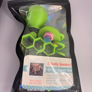 May include: A clear plastic bag containing a green spider-shaped toy, a green honeycomb-shaped object, and a business card. The business card has a photo of a spider and the text "Spindly Spooders".