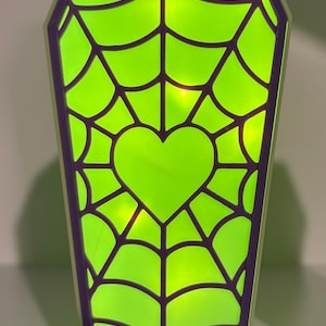 Puede incluir: Una lámpara con forma de ataúd que presenta un centro iluminado en verde lima vibrante con una superposición de telaraña morada y una forma de corazón sólido en el centro. El marco es de color claro y emite un brillo intenso desde el interior.