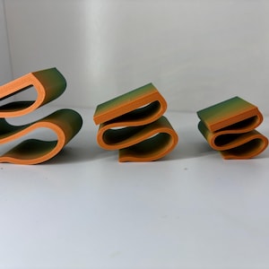 May include: Three 3D printed objects arranged in descending order of size. Each object features a continuous, folded ribbon-like structure with a gradient colour transition from orange at the base to a muted green at the top edge.