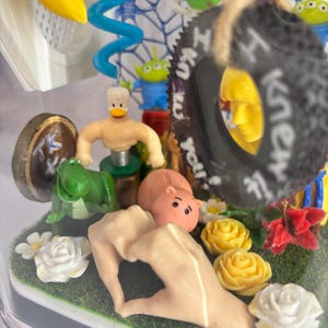 May include: A clear display case containing a collection of miniature figures including a green dinosaur, a pink hippo, a duck-headed figure, and a black tire ornament with the text I knew it. The scene features artificial grass and flowers.