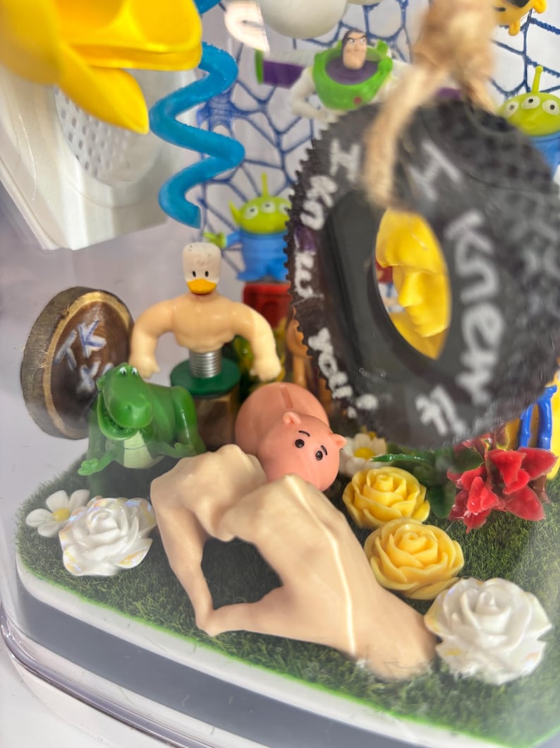 May include: A clear display case containing a collection of miniature figures including a green dinosaur, a pink hippo, a duck-headed figure, and a black tire ornament with the text I knew it. The scene features artificial grass and flowers.