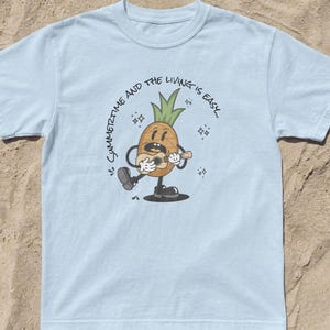 Può includere: T-shirt azzurra con un ananas dei cartoni animati che suona l'ukulele. L'ananas ha una faccia sorridente e indossa le scarpe. Il testo "Summertime and the living is easy..." è stampato intorno all'ananas.
