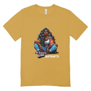 Op de afbeelding: Mosterdgeel T-shirt met een kleurrijke illustratie van een persoon met donker haar, een jas, jeans en sneakers. De tekst "UNAPOLOGETIC" staat eronder. Het shirt is gemaakt van een zacht materiaal.
