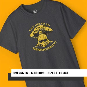 Puede incluir: Camiseta gris oscuro con un gráfico amarillo de un casco y una nube en forma de hongo, con el texto "SAY HELLO TO DEMOCRACY!". La camiseta también tiene el texto "OVERSIZES - 5 COLORS - SIZES L TO 3XL".