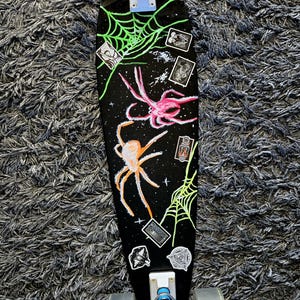 Könnte beinhalten: Schwarzes Longboard-Skateboard mit Spinnen- und Spinnennetz-Design. Das Board hat einen schwarzen Hintergrund mit neongrünen Spinnennetzen und rosa und orangefarbenen Spinnen. Mehrere Aufkleber sind ebenfalls auf dem Board.