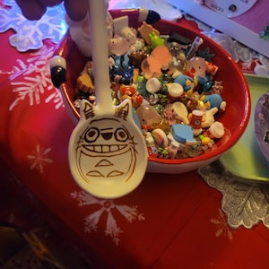 May include: A white ceramic spoon with a cartoon character design, held in front of a red bowl filled with small toys. The spoon features a smiling face with large eyes and a wide grin. The bowl sits on a red tablecloth with white snowflake patterns.