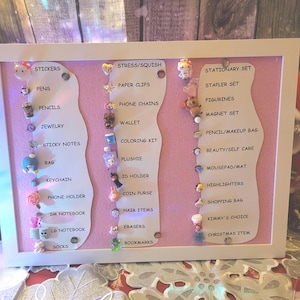 May include: A framed display board with a pink background and white frame. The board features various small items like stickers, pens, pencils, and keychains. Text labels categorize the items, including "Stickers," "Pens," and "Jewellery."